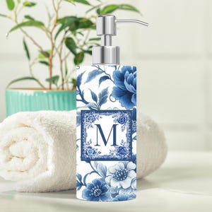 May include: A rectangular soap dispenser with a stainless steel pump. The dispenser is decorated with a blue floral pattern on a white background. A blue framed letter "M" is centered on the front. The dispenser is on a white surface.