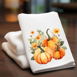 Puede incluir: Toalla de cocina blanca con un diseño de acuarela de calabazas naranjas, flores amarillas y hojas verdes. La toalla está doblada y apilada con otras dos toallas blancas. El diseño está centrado en la toalla.