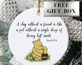 May include: White ceramic ornament featuring Winnie the Pooh sitting with a pot of honey. The ornament has the quote "A day without a friend is like a pot without a single drop of honey left inside" and is attributed to Winnie the Pooh.