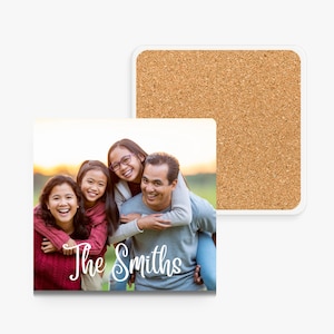 A square photo coaster with a white border. The coaster features a family of four smiling and looking at the camera. The family is standing in a field with a sunset in the background. The text 'The Smiths' is written in white cursive font below the family.