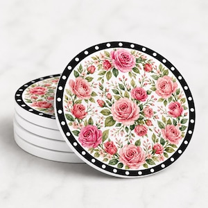 May include: A stack of round ceramic coasters featuring a white background with a floral pattern of pink roses, green leaves, and small buds. Each coaster has a black border decorated with a repeating pattern of small white dots around the edge.
