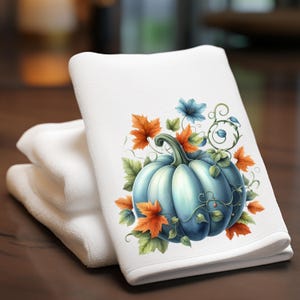 May include: White hand towel with a light blue pumpkin design, surrounded by orange and green leaves and blue flowers. The towel is folded and stacked with two other white towels. The design is centered on the towel.