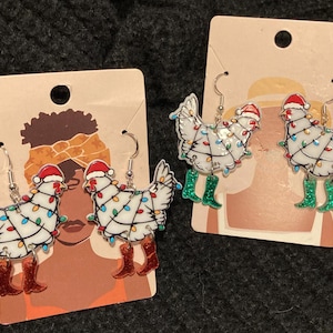 Könnte beinhalten: Ein Paar Hängeohrringe mit weißen Hühnern mit roten Weihnachtsmannmützen und grünen Stiefeln, verziert mit weißen Weihnachtslichtern. Die Ohrringe befinden sich auf einer Karte mit einer Zeichnung einer Person mit einem braunen Stirnband.