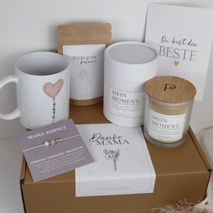 Könnte beinhalten: Eine Geschenkbox mit einer weißen Tasse mit einem rosa Herzballon-Design und dem Wort "mama". Die Box enthält auch eine Kerze, ein Armband, eine Karte mit Text und einen Teebeutel. Die Box ist braun mit weißen Etiketten.