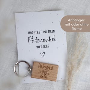 Könnte beinhalten: Ein Geschenkset mit einer Karte mit dem Text "Möchtest du mein Patenonkel werden?" und einem Holzschlüsselanhänger. Der Schlüsselanhänger ist mit "Patenonkel von Leo" graviert. Das Set enthält einen dekorativen Zweig aus getrocknetem Gras.