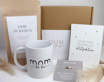 May include: A gift set including a brown paper pouch labeled LIEBE IM BAUCH, a white ceramic mug with the text mom to be, a card reading MOM TO BE, a card with the text GROBE DINGE BEGINNEN OFT klitzeklein, and a bracelet on a card labeled MOM TO BE.