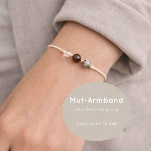 Könnte beinhalten: Zartes Armband mit cremefarbenem Band und drei dekorativen Perlen. Die Perlen umfassen einen klaren Kristall, eine dunkelbraune Perle und einen gesprenkelten Stein. Der Text "Mut-Armband" ist sichtbar.
