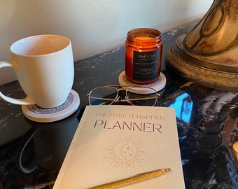 Könnte beinhalten: Ein beiger Planer mit dem Text "The Make It Happen Planner" und einem goldenen Stift darauf. Der Planer hat ein Mond- und Sternen-Design und den Text "Daily Summary of My To Do's | Priorities | Goals & Reminders".