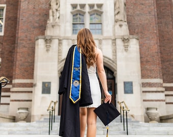 May include: A person wearing a white dress and white high-heeled sandals, carrying a black graduation gown with a blue and gold sash that reads Class of 2021, and holding a black graduation cap with a blue and gold tassel while walking up stairs.