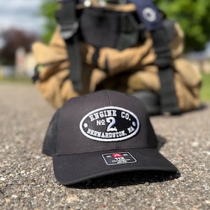 May include: Black trucker hat with a white patch that reads "ENGINE CO. NO. 2 BERNARDSTON, MA." The hat is sitting on a textured surface, with a blurred background of firefighting gear.