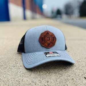 May include: A gray and black trucker hat with a brown leather patch featuring the text "International Association of Fire Fighters Local 12" and a fire hydrant graphic.