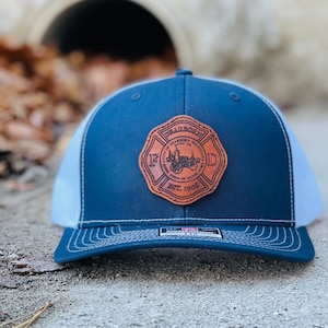 May include: A navy blue and white trucker hat with a leather patch. The patch features the text "Dearborn Michigan" and "Home Town of Henry Ford" with a fire department emblem. The hat has a curved brim and a mesh back.