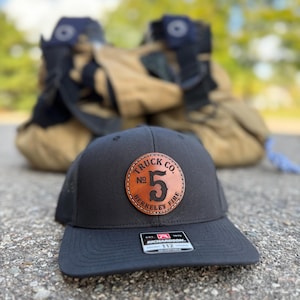 May include: Black baseball cap with a leather patch that reads "Truck Co. No. 5 Berkeley Fire." The cap is sitting on a concrete surface, with firefighting gear in the background.