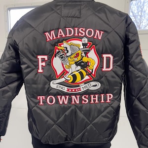 May include: Black quilted bomber jacket with a diamond pattern. The back features an embroidered emblem with a cartoon bee holding a fire hose, the words "MADISON F D TOWNSHIP" in red, and "ENG XXXI MED".