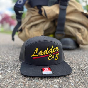 May include: A black snapback hat with a flat brim and mesh back panels. The front features embroidered yellow text that reads Ladder Co.5 with a red horizontal line underneath. A rectangular sticker is attached to the center of the brim.
