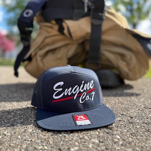 May include: A navy blue snapback cap with a flat bill and mesh back panels. The front features white embroidered text that reads Engine Co.7 with a thin red horizontal line underneath. A rectangular sticker is attached to the top of the bill.