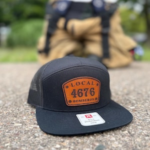 May include: A black trucker hat with a flat brim and a brown leather patch on the front. The patch features the text LOCAL 4676 BOMBEROS. A white sticker on the brim displays the Richardson brand logo and the text It's a Richardson 863M.