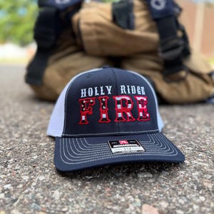 May include: A navy blue and white trucker hat with the text "HOLLY RIDGE FIRE" in red and white. The hat has a curved brim and a mesh back. A firefighter's uniform is in the background.
