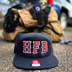 May include: A navy blue flat-brimmed cap with the letters "HFD" in red, outlined in white. The cap has a tag from the brand "Flat Bill Snapback". Firefighting gear is blurred in the background.