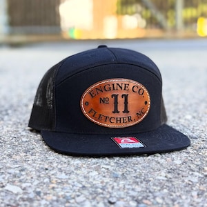 May include: Black trucker hat with a curved brim and mesh back. The front features a brown leather patch with the text "ENGINE CO. NO 11 FLETCHER, NC" in a circular design. The hat has a flat bill.