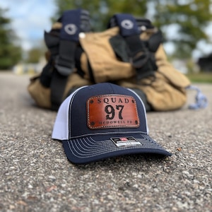 May include: A navy blue and white trucker hat with a leather patch that reads "SQUAD 97 MCDOWELL FD." The hat is sitting on a textured surface, with a blurred background of firefighting gear.