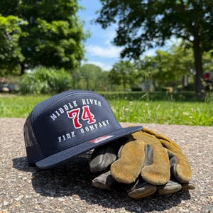 May include: A navy blue trucker hat with a mesh back and a flat brim. The front features white embroidered text reading MIDDLE RIVER FIRE COMPANY with the number 74 in red. Beside the hat sits a pair of worn, tan-colored leather work gloves.