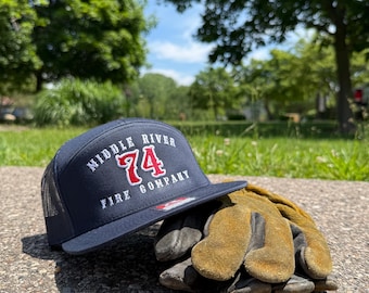 May include: A navy blue trucker hat with a mesh back and a flat brim. The front features white embroidered text reading MIDDLE RIVER FIRE COMPANY with the number 74 in red. Beside the hat sits a pair of worn, tan-colored leather work gloves.