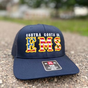 May include: Navy blue baseball cap with the text "CONTRA COSTA CO." above the letters "RMS" in red, white, and blue. The hat has a curved brim and a mesh back.