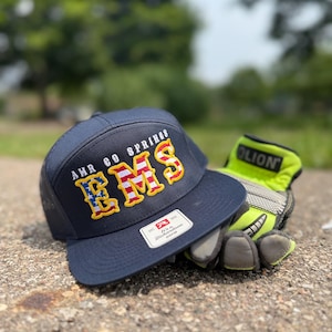 May include: A navy blue baseball cap with a mesh back and a flat brim. The front features embroidered text reading AMR CO SPRINGS and EMS, with the letters EMS filled with a red, white, and blue American flag pattern and outlined in yellow.