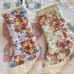 Op de afbeelding: Twee gewatteerde kerstsokken met bloemenpatchworkpatronen in rood, geel en blauw. De sokken hebben een crèmekleurige manchet en teen, en een lus om op te hangen. Ze worden getoond op een vintage quilt.