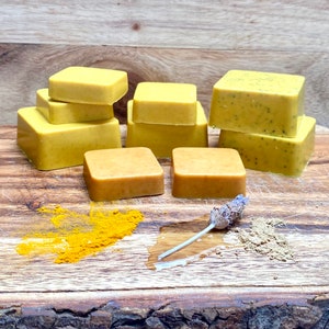 May include: A wooden surface with six bars of yellow soap, a small pile of turmeric powder, a sprig of lavender, and a small pile of brown powder.