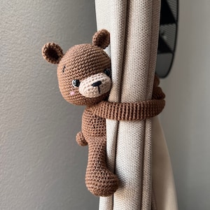 May include: A brown crocheted teddy bear curtain tieback. The bear is holding onto a beige curtain with its arms.