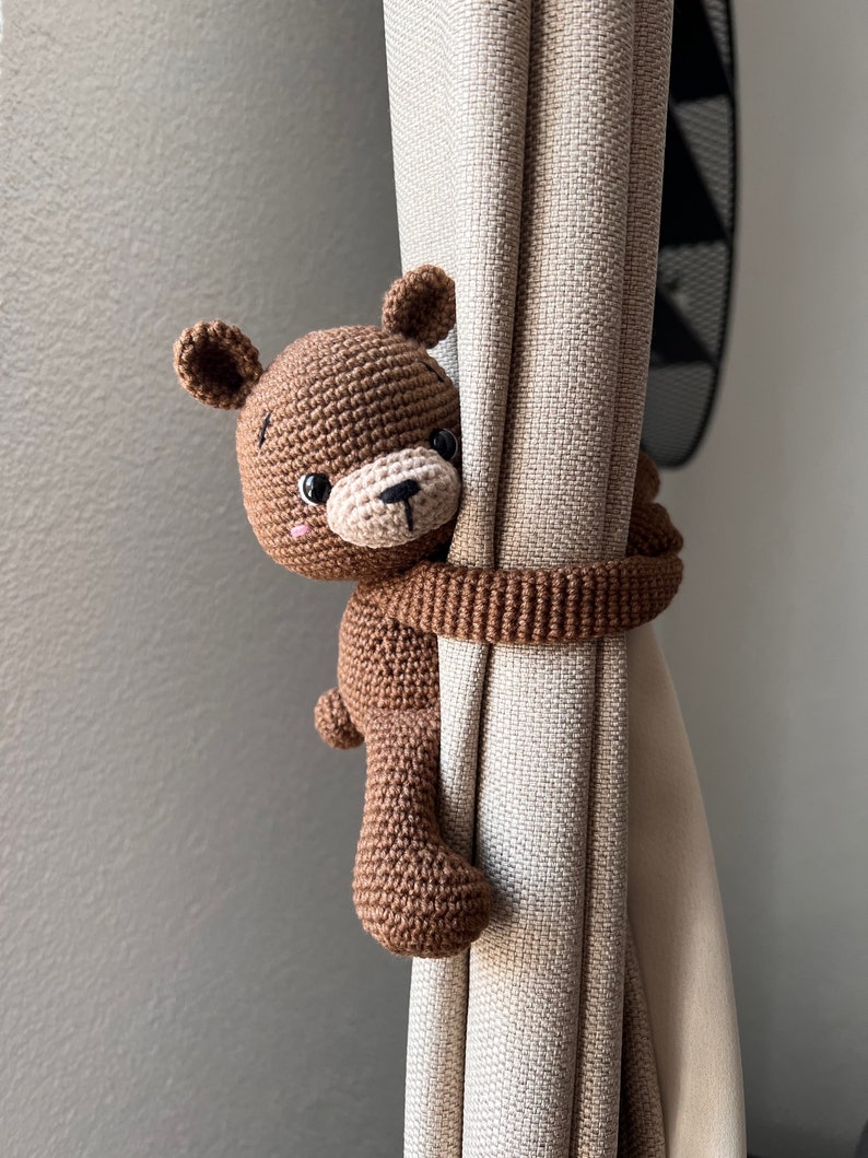 May include: A brown crocheted teddy bear curtain tieback. The bear is holding onto a beige curtain with its arms.