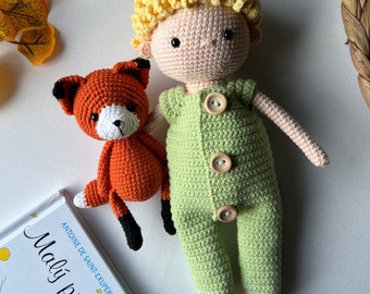 May include: A crocheted plush toy of The Little Prince, a boy with blonde hair, wearing a green jumpsuit and holding a crocheted fox toy. The book "The Little Prince" by Antoine de Saint-Exupéry is in the background.