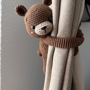 May include: A brown crocheted bear holding a beige curtain. The bear has black eyes and a black nose.