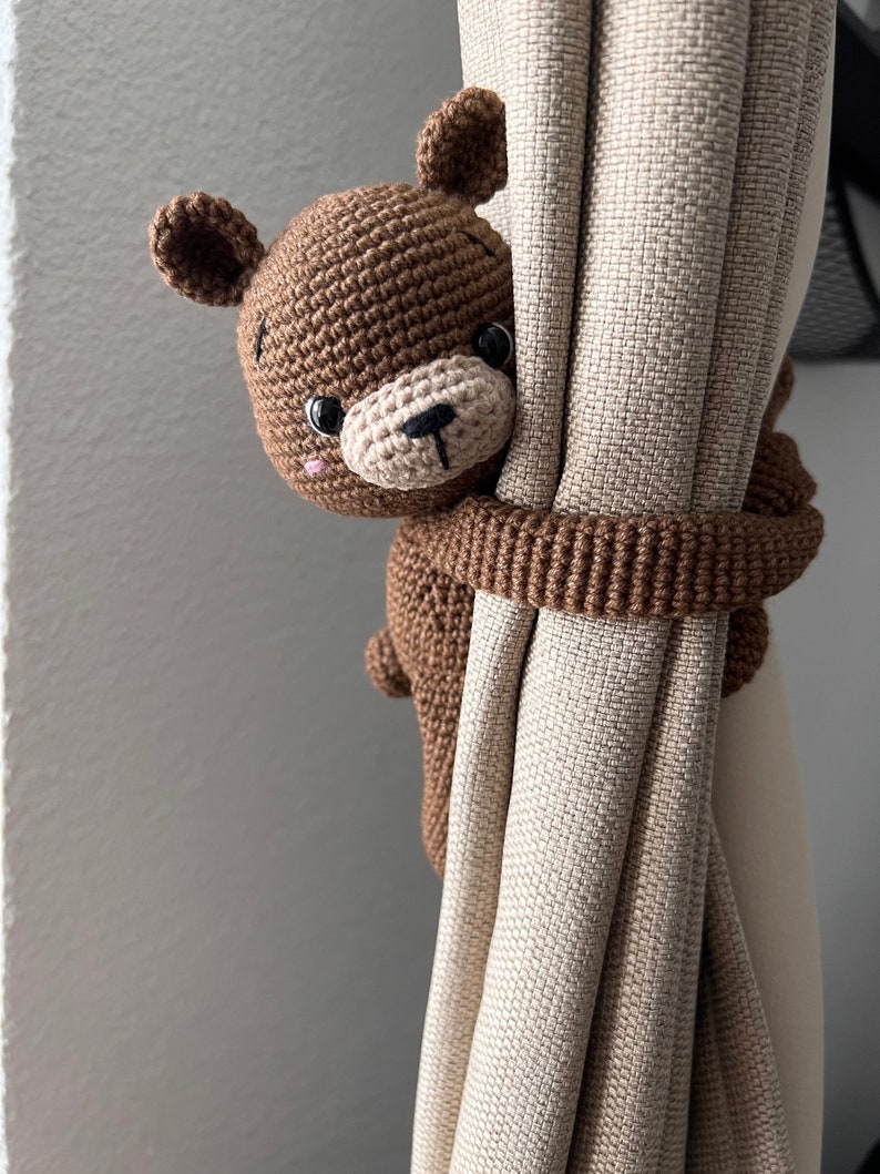 May include: A brown crocheted bear holding a beige curtain. The bear has black eyes and a black nose.