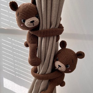 May include: Two brown crocheted teddy bears are used as curtain tiebacks. The bears are holding onto a beige curtain with their arms.