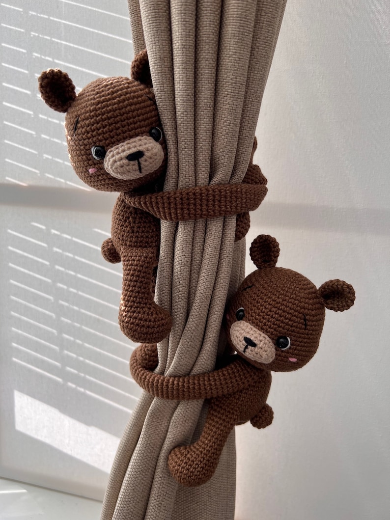 May include: Two brown crocheted teddy bears are used as curtain tiebacks. The bears are holding onto a beige curtain with their arms.