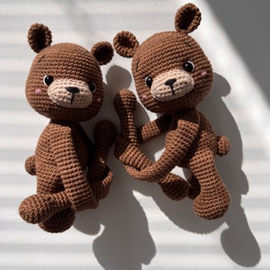 May include: Two brown crocheted teddy bears with black eyes and pink cheeks. The bears are sitting on a white surface.