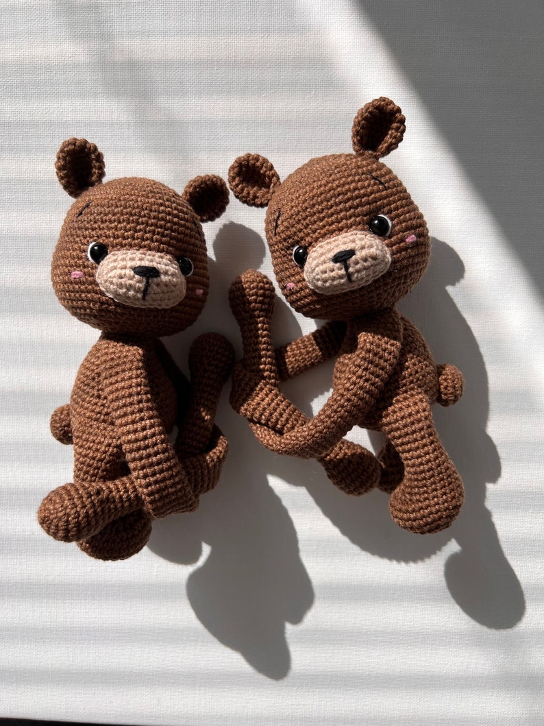 May include: Two brown crocheted teddy bears with black eyes and pink cheeks. The bears are sitting on a white surface.