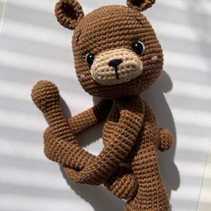 May include: A brown crocheted teddy bear with black eyes and a beige nose. The bear is sitting with its legs crossed and its arms outstretched.