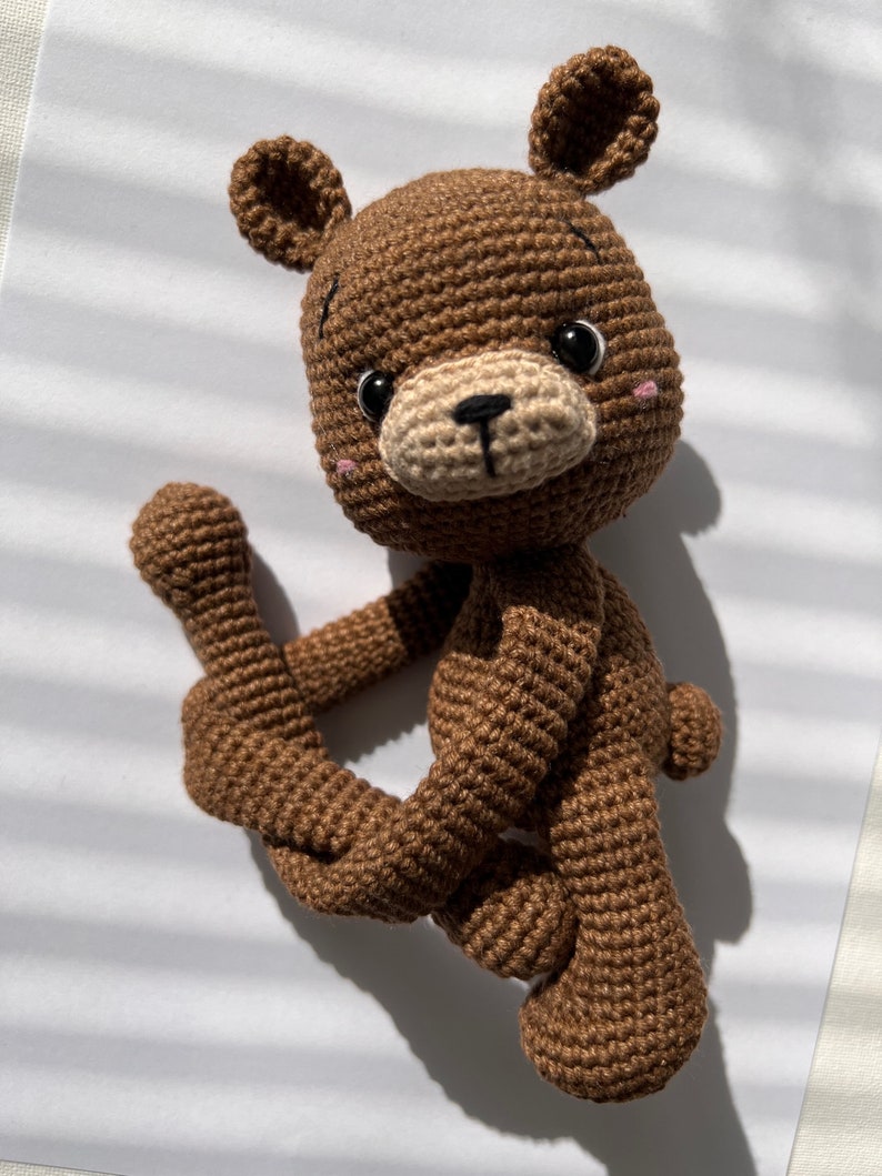 May include: A brown crocheted teddy bear with black eyes and a beige nose. The bear is sitting with its legs crossed and its arms outstretched.