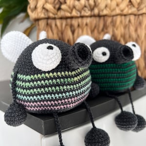 May include: Two crocheted amigurumi toys shaped like bugs with black bodies, white eyes, and long legs. One bug has green and pink stripes and the other has green stripes.
