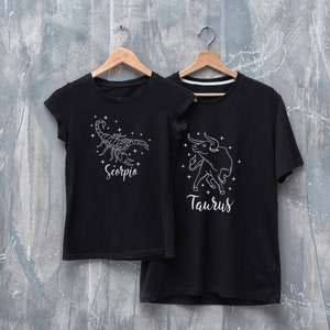 May include: Two black t-shirts with white zodiac signs. The left shirt has a scorpion with the word "Scorpio" underneath. The right shirt has a bull with the word "Taurus" underneath.