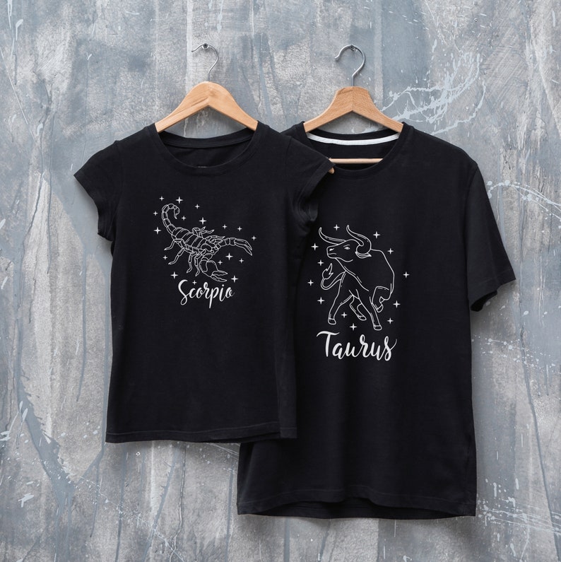 May include: Two black t-shirts with white zodiac signs. The left shirt has a scorpion with the word "Scorpio" underneath. The right shirt has a bull with the word "Taurus" underneath.
