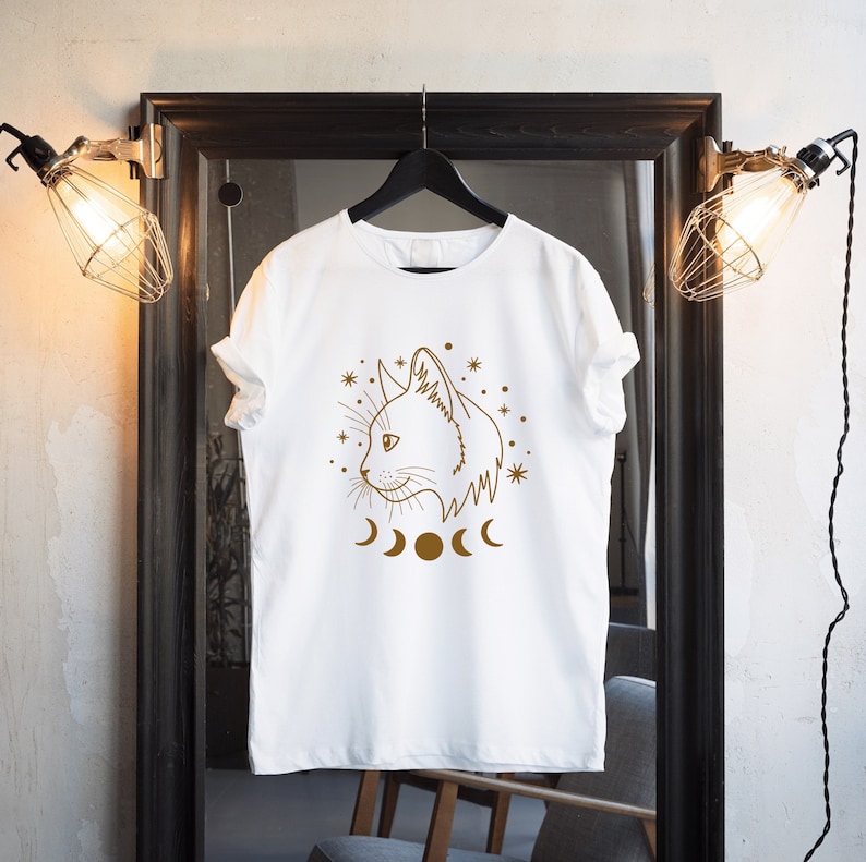 May include: White t-shirt with a brown line drawing of a cat's face with a crescent moon and stars around it.