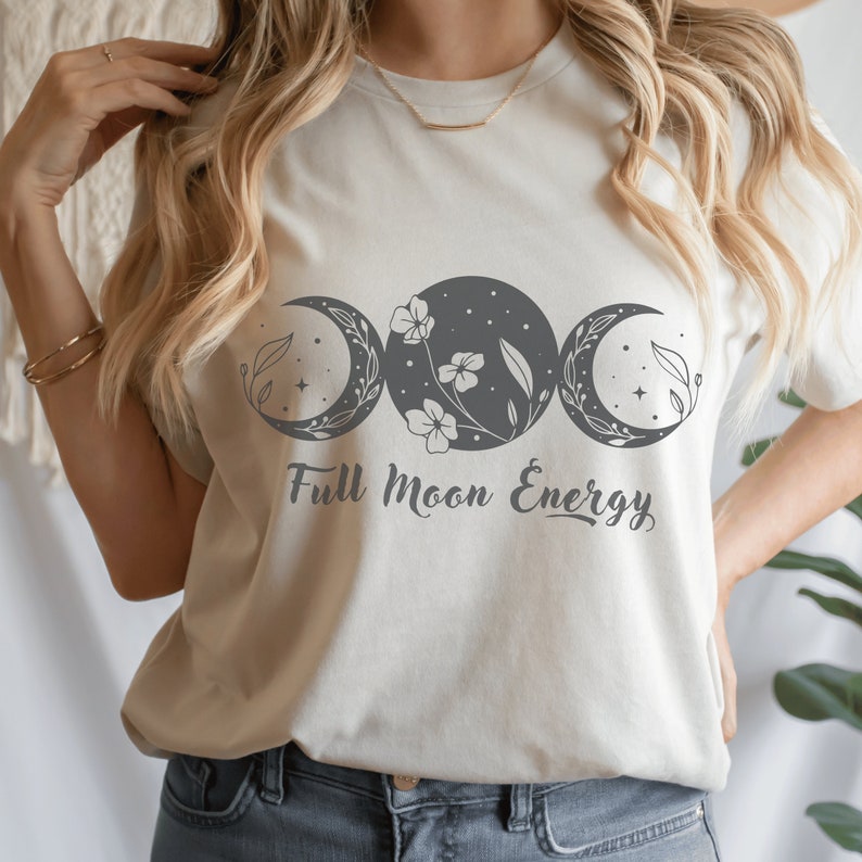 Op de afbeelding: Een wit T-shirt met een grijze afbeelding van drie manen met bloemen en bladeren. De tekst "Full Moon Energy" is onder de manen gedrukt.
