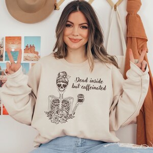 May include: A beige sweatshirt with a graphic of a skeleton holding a coffee cup. The skeleton has a bun in its hair and heart-shaped sunglasses. The text on the sweatshirt reads "Dead inside but caffeinated."