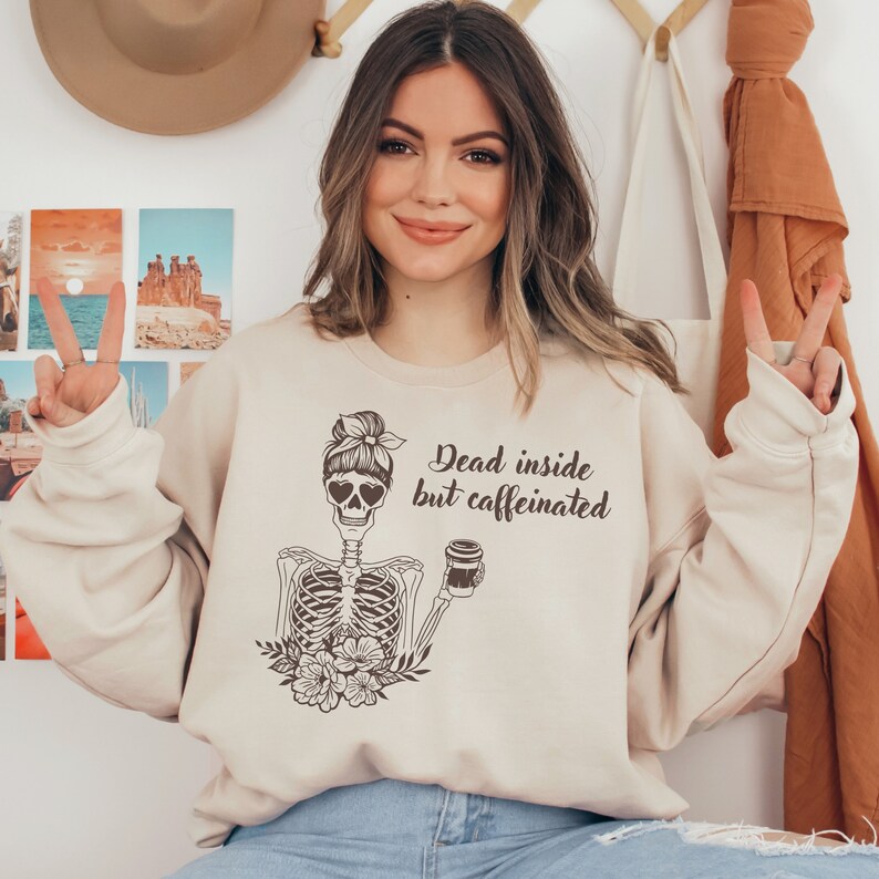 May include: A beige sweatshirt with a graphic of a skeleton holding a coffee cup. The skeleton has a bun in its hair and heart-shaped sunglasses. The text on the sweatshirt reads "Dead inside but caffeinated."