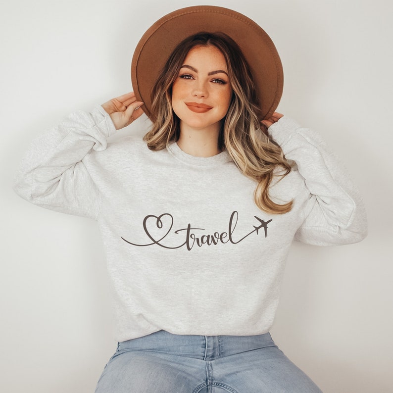 Puede incluir: Una sudadera gris con un coraz&oacute;n marr&oacute;n y un gr&aacute;fico de avi&oacute;n y el texto "travel" en cursiva.
